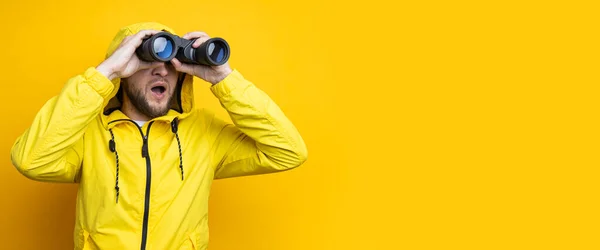 Surprised young man in yellow raincoat looking through binoculars on yellow background. Banner.