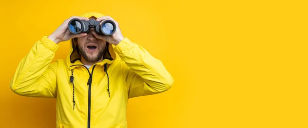Surprised young man in yellow raincoat looking through binoculars on yellow background. Banner.