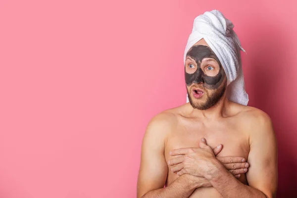surprised naked man with a cosmetic mask on his face, covering his chest with his hands on a pink background.