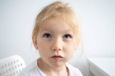 Child girl blonde sits on a white chair at the table on a white background.