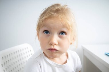 Child girl blonde sits on a white chair at the table on a white background.