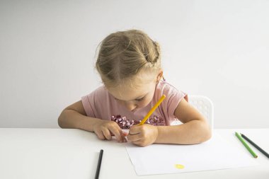 The child girl draws with pencils sitting at a white table on a white background.