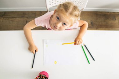 The child girl draws with colored pencils sitting at a white table. Top view, flat lay.