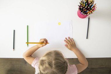 The girl draws with colored pencils sitting at a white table. Top view, flat lay.