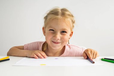 A smiling child blonde girl draws with colored pencils sitting at a white table.
