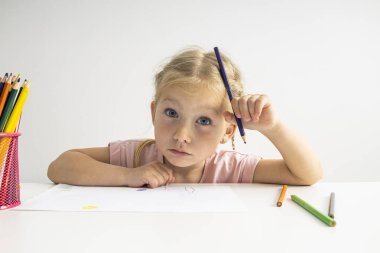 Blue-eyed blonde child girl draws with colored pencils sitting at a white table.