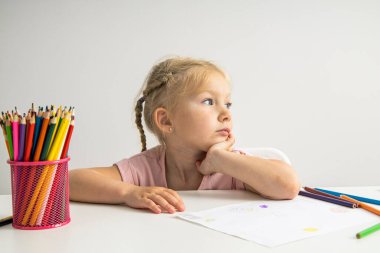 A thoughtful child blonde girl draws with colored pencils sitting at a white table.