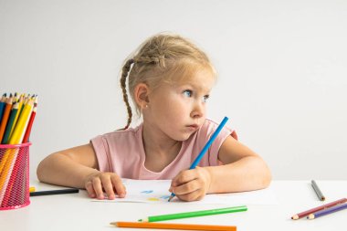 A blonde child girl draws with colored pencils sitting at a white table.
