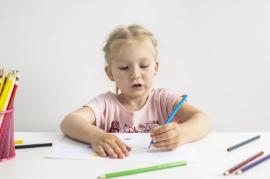 Blonde child girl draws with colored pencils sitting at the table.