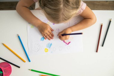 Blonde child girl draws with colored pencils sitting at the table. Top view, flat lay.