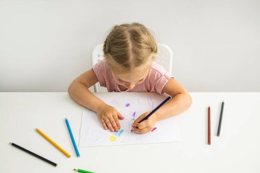 Blonde child girl draws with colored pencils sitting at the table. Top view, flat lay.