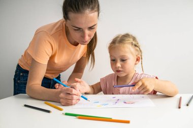 Mom helps her daughter draw with colored pencils on paper at the table.
