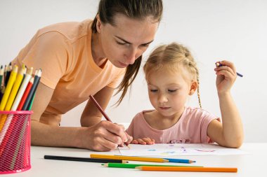 The kindergarten teacher helps the child girl to draw with colored pencils on paper.