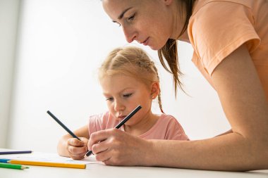 The kindergarten teacher helps the child girl to draw with colored pencils on paper.