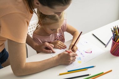 Mom and daughter draw together with colored pencils on paper at the table.