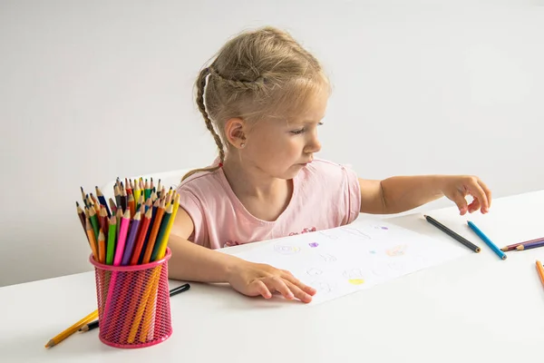 A blonde child girl draws with colored pencils sitting at a white table.