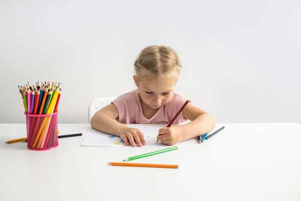 Blonde child girl draws with colored pencils sitting at the table.