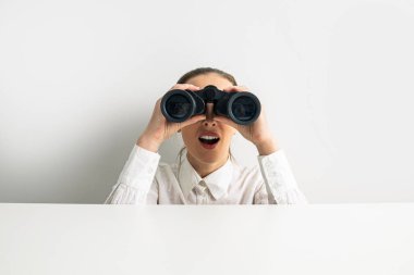 Surprised woman looking through binoculars peeking out from behind white table.