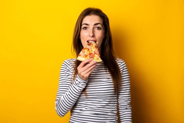 young woman bites a slice of hot fresh pizza on a yellow background.