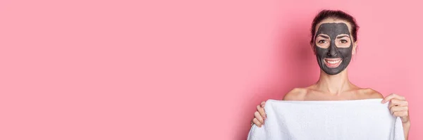 nude smiling girl hides behind a towel with a clay mask on her face on a pink background. Banner