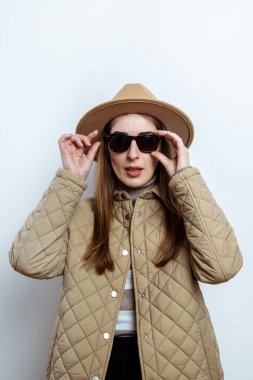 Beyaz bir duvara karşı güneş gözlüklü ceketli genç bir kadın.