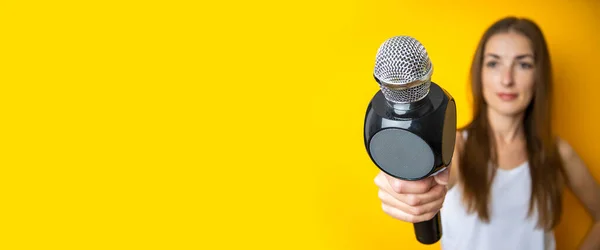Young woman holding a microphone, interviewing on a yellow background. Reporter or journalist concept. Banner.