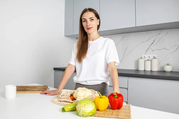 Smiling young woman standing near table with fresh vegetables in kitchen.