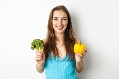 Elinde brokoli ve beyaz arka planda yeşil biber tutan gülümseyen genç bir kadın..