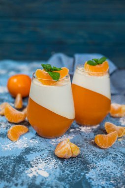 Mandalinalı cam bardakta tatlı jöleli puding ve mavi arka planda nane yaprağı.
