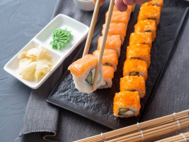 Philadelphia ve California desenli siyah bir tabakta yuvarlanıyor. Bir rulo, diğerlerinin üstündeki büyükanne çubuklarında tutuluyor. Wasabi ve zencefilin yanında. Yan görüş. Japon mutfağı.