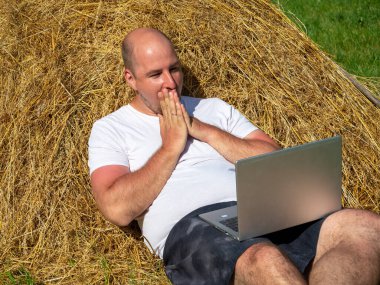 Orta yaşlı bir adam, elinde gri bir dizüstü bilgisayarla sarı bir samanlıkta yatıyor. Uzaktan çalışma, eğitim. Kırsal bölge. Yüzünde şaşırmış bir ifade, elleri ağzını kapatıyor.