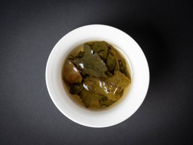 Siyah arka planda demlenmiş çay yaprakları olan beyaz bir kase. Yaklaş, yan görüş. Çin çayı, açık yapraklar. Üst manzara, düz uzanma