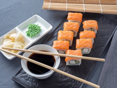 Suşi Philadelphia 'yı gri arka planda siyah desenli bir tabakta yuvarlıyor. Wasabi zencefili ve sos çubukları. Somon, krem peynir, avokado..