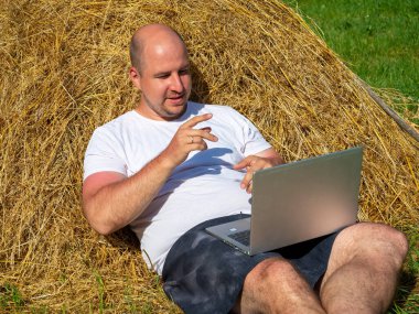 Tişört ve şort giymiş orta yaşlı bir adam elinde gri bir dizüstü bilgisayarla sarı bir samanlıkta yatıyor. Uzaktan çalışma, eğitim. Kırsal bölge. Müdahaleciye bir el hareketi gösterir