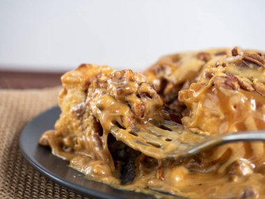 Karamel serpilmiş ve fındıkla süslenmiş tarçınlı çöreğin yakın çekimi siyah bir tabakta ve keten peçetede yatıyor. Yan manzara, bir parça tatlı çatalla alınır. Seçici odak