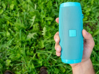 Mavi kablosuz müzik hoparlörünü tutan bir adamın yakın çekimi. Arka planda kalın yeşil bir çim var. Açık hava aygıtı kullanarak. Üst Görünüm, Boşluğu Kopyala