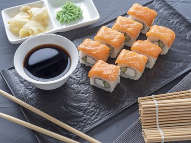 Suşi Philadelphia 'yı gri arka planda siyah desenli bir tabakta yuvarlıyor. Wasabi zencefili ve sos çubukları. Somon, krem peynir, avokado..