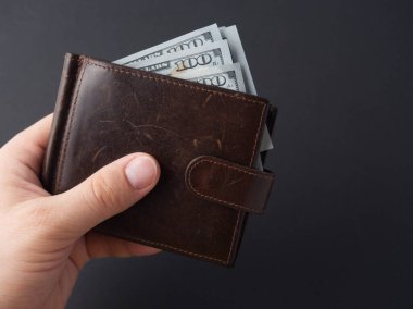 Bir adamın elinde siyah arka planda 100 dolarlık banknotlar olan kahverengi deri bir cüzdan var. Para, zenginlik, zenginlik kavramı