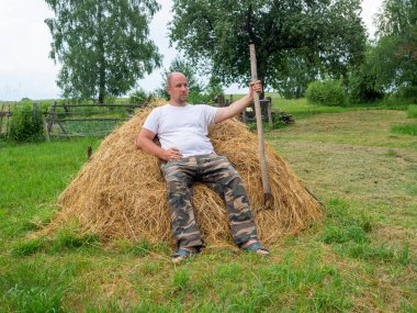 Pantolon ve tişört giymiş orta yaşlı bir adam sarı saman yığınının üzerinde oturuyor ve elinde bir yaba tutuyor. Kırsal bölgeler ve tarım işçileri kavramı