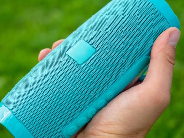 Mavi kablosuz müzik hoparlörünü tutan bir adamın yakın çekimi. Yeşil bulanık arkaplan