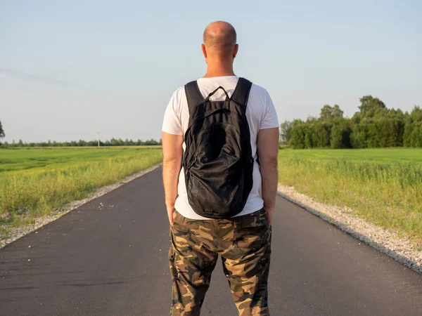 A man with a backpack on his back stands with his back to the camera on an empty asphalt road during the day. The concept of travel and hiking, adventure, vacation.