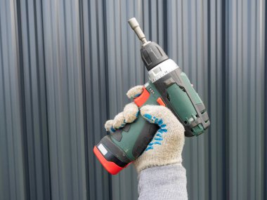 Elinde kablosuz tornavida tutan bir adamın yakın çekimi. Arka planda gri metal bir çit var. Çitlerin inşa ve montajı konsepti. Mobil araç