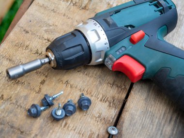 Tahta bir arka planda, üzerinde hortumu olan elektrikli bir tornavidayı yakından çek. Onun yanında, metal üzerine çatı vidaları dizilmiştir. Tamir ve inşaat kavramı