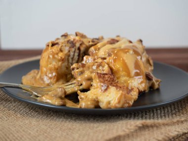 Karamel serpilmiş ve fındıkla süslenmiş tarçınlı çöreğin yakın çekimi siyah bir tabakta ve keten peçetede yatıyor. Yan manzara, bir parça tatlı çatalla alınır. Seçici odak