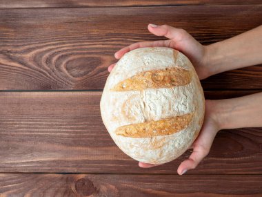 Bir somun buğday ekmeğini tutan ellerin yakın çekimi. Kahverengi ahşap arka plan, Üst Manzara, düz yatış. Fırın kavramı