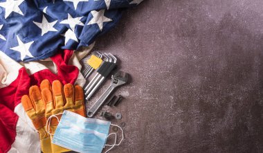 Coronavirus hastalığı (Covid-19) sırasındaki İşçi Bayramı konseptin kutlu olsun. Koyu taş arka planda farklı inşaat araçları ve tıbbi maskeli Amerikan bayrağı.