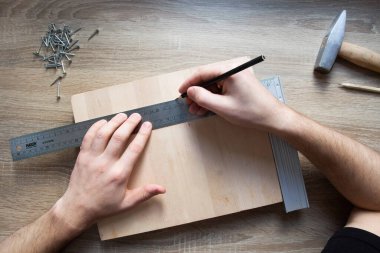 Kendin yap: çocuk eğitim oyunu - karanfilli tahta. 2. adım, tahta zeminde dikdörtgen bir tahta var ve elleri metal bir cetvel tutuyor ve tahtadaki delikleri işaretliyor. Yakınlarda çivi ve çekiç var.