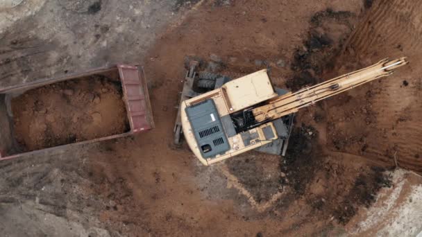 Yellow hydraulic excavator digs earth for foundation of house at a construction site and loads earth into truck, aerial top view — Stock Video