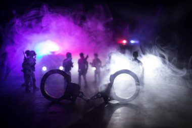 Gece polis baskını ve sen tutuklusun. Arka tarafında polis arabası olan kelepçelerin silueti. Görüntü sisli arka planda yanıp sönen kırmızı ve mavi polis ışıkları. Seçici odak