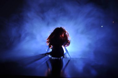 Hafif arka planda bir kadının silueti. Gece karanlığında uzun saçlı, sisli arka planda mavi ışıkları olan yalnız bir oyuncak bebek. Seçici odak